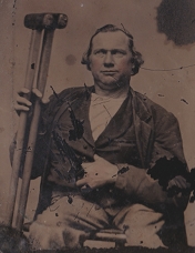 This is an image of a man seated. He has a few of his left fingers tucked into his jacket. He is hold a pair of crutches at right.