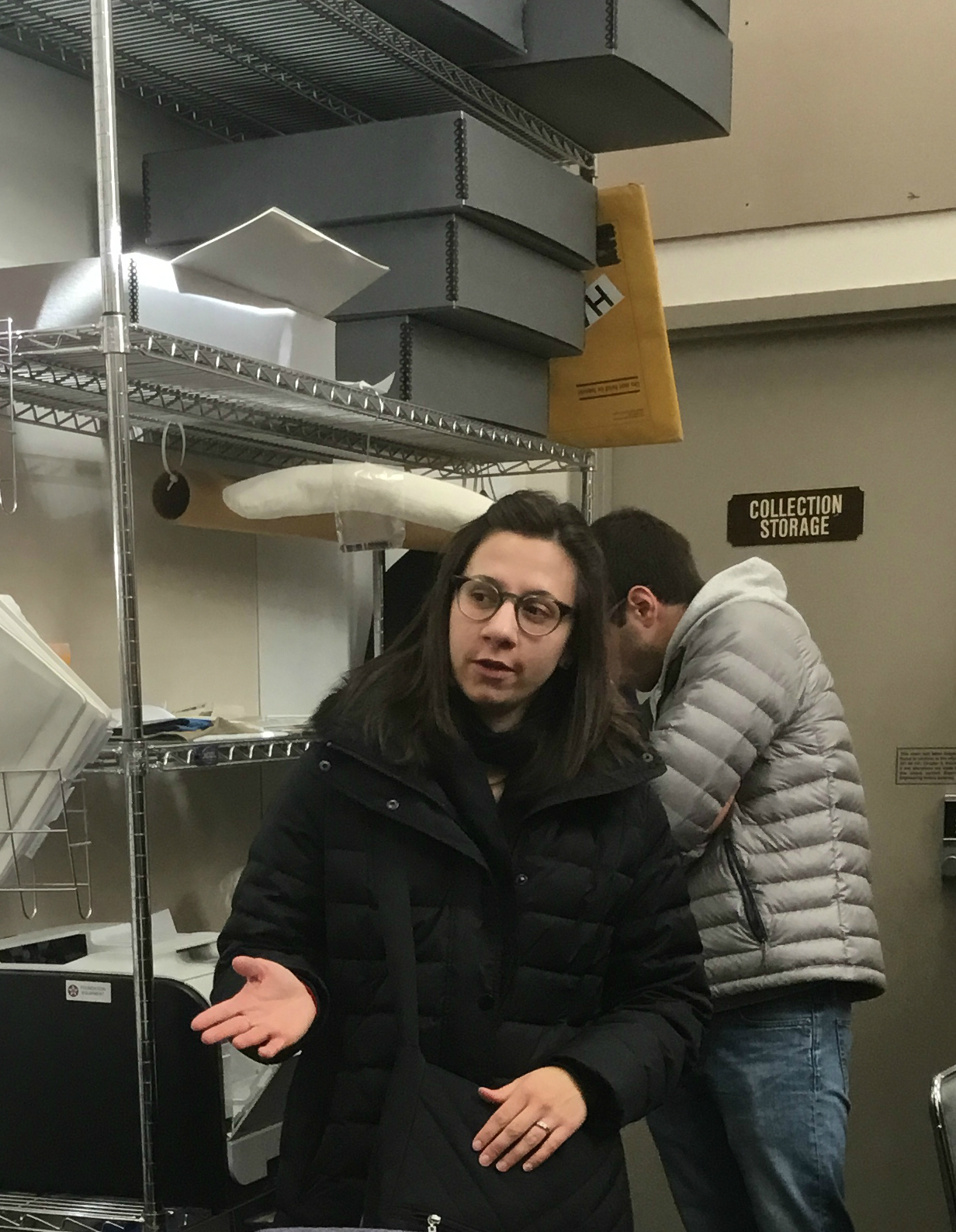 This is a photograph of me standing an gesturing with my right hand. There is a student (face obscured) standing behind me. I am in front of a large metal shelf that contains gray archival boxes.