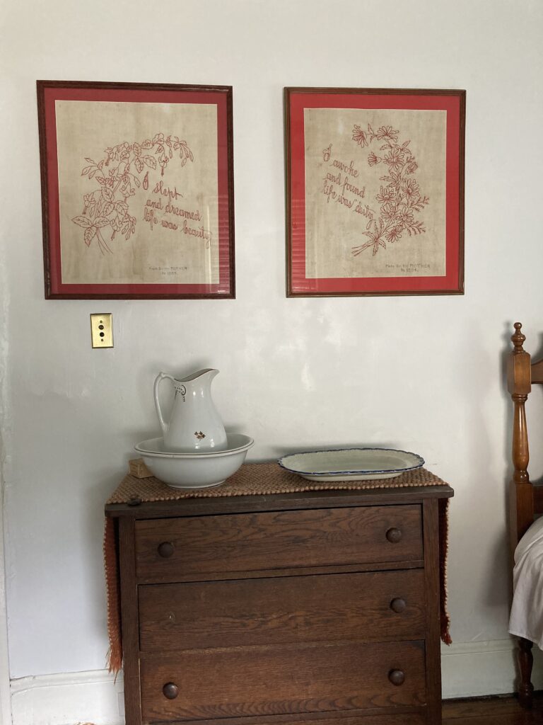This is a color photograph of two pieces of framed needlework on a white wall. They are having above a dresser that has a ceramic wash bowl and basin on it as well as blue and white platter.